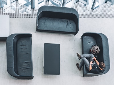 An overhead view of a modern waiting area with three black leather sofas arranged around a rectangular black table on a light gray floor. A person is seated on the right sofa, looking at a phone, with a backpack placed beside them.