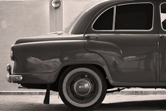 A sepia-toned image of a vintage car showing the rear half, highlighting the classic curved fender and large round taillight. The car is parked on a tiled pavement with a textured wall and a sconce lamp in the background. The windows of the car are tinted, and the hubcap is noticeably reflective.