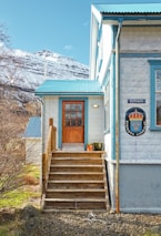 A cozy house located in a picturesque countryside setting. The building features a light-colored stone exterior with blue trim and a wooden staircase leading up to a door with a window. A coat of arms and a sign with text are visible on the house wall. In the background, snow-capped mountains rise under a clear sky. Bare trees and green grass surround the house, with a couple of potted plants near the entrance.