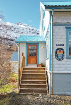 A cozy house located in a picturesque countryside setting. The building features a light-colored stone exterior with blue trim and a wooden staircase leading up to a door with a window. A coat of arms and a sign with text are visible on the house wall. In the background, snow-capped mountains rise under a clear sky. Bare trees and green grass surround the house, with a couple of potted plants near the entrance.
