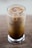 A tall glass filled with iced coffee, showcasing a gradient of creamy beige and rich brown hues. Large ice cubes are visible on the surface, creating a refreshing appearance.