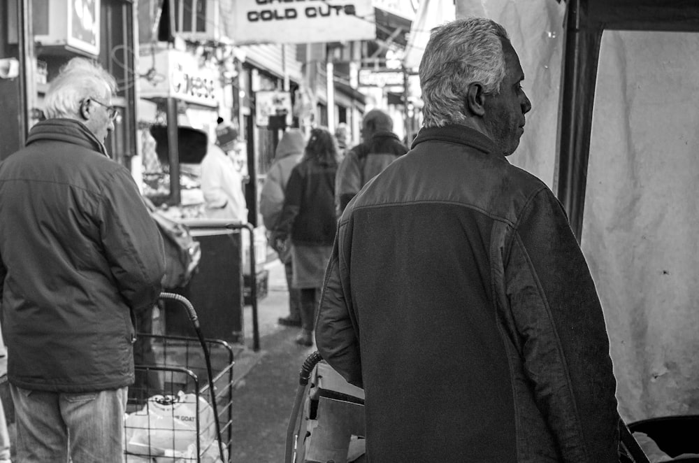 man in black jacket standing near people walking on street in grayscale photography