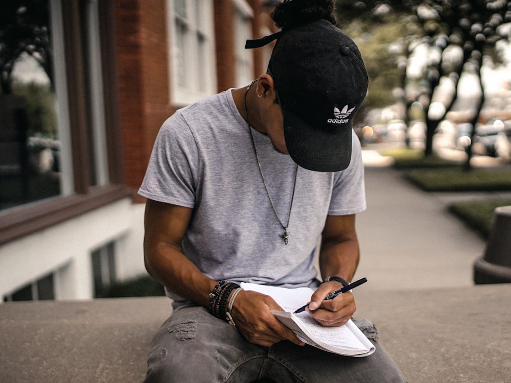 pessoa de boné adidas preto sentado no banco escrevendo no caderno