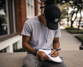 person in black adidas cap sitting on bench writing on notebook