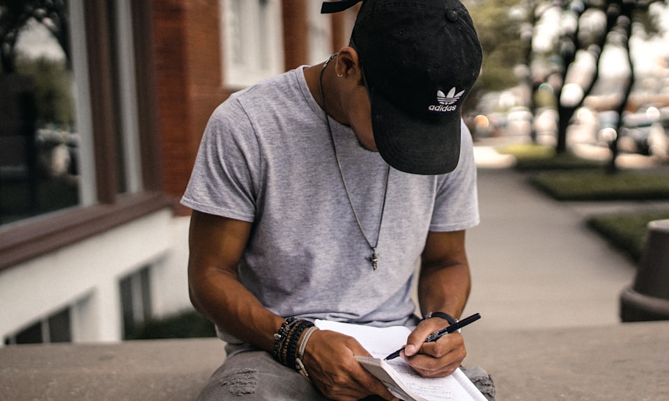 person in black adidas cap sitting on bench writing on notebook