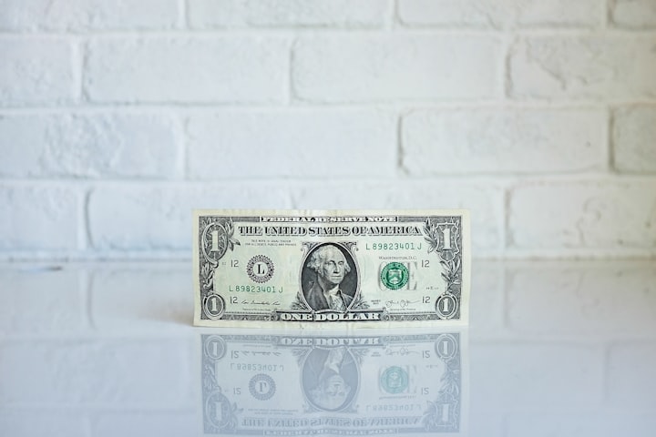 An dollar bill upright on a white surface with a white brick wall in the background 