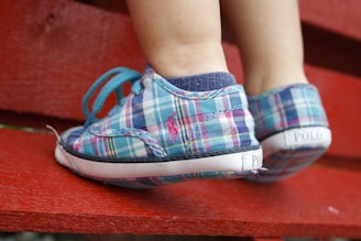 Close-up of a child's feet wearing colorful plaid sneakers with teal laces. The child is standing on a bright red wooden surface.