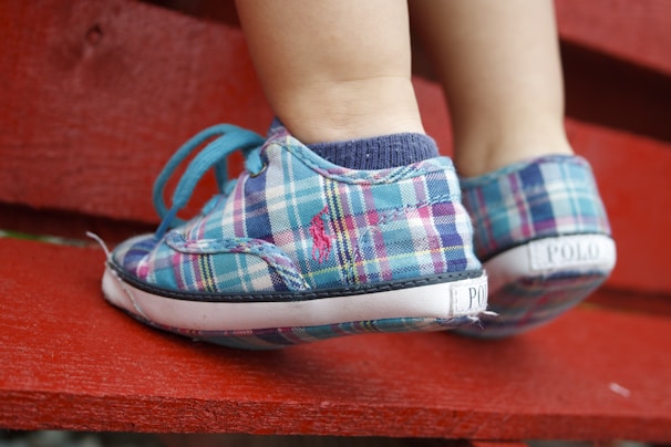 Close-up of a child's feet wearing colorful plaid sneakers with teal laces. The child is standing on a bright red wooden surface.