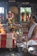 A man dressed in traditional attire is performing a Hindu ritual. He is seated beside a small altar adorned with colorful flowers and garlands. In front of him, there is a fire burning. Several religious statues are visible in the background, also decorated with floral garlands.