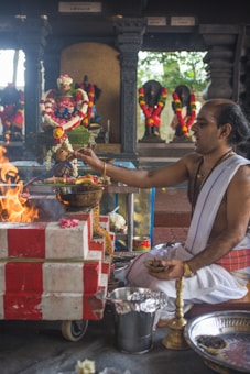 A man dressed in traditional attire is performing a Hindu ritual. He is seated beside a small altar adorned with colorful flowers and garlands. In front of him, there is a fire burning. Several religious statues are visible in the background, also decorated with floral garlands.