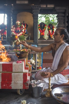 A man dressed in traditional attire is performing a Hindu ritual. He is seated beside a small altar adorned with colorful flowers and garlands. In front of him, there is a fire burning. Several religious statues are visible in the background, also decorated with floral garlands.