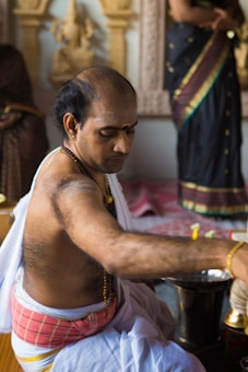 A man dressed in traditional attire is engaged in a ritual or ceremony, surrounded by ornate decorations. He wears a red and white dhoti, jewelry, and has a sacred thread across his body. Another person in a saree is partially visible in the background.