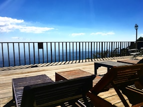 A wooden deck with lounge chairs overlooks a vast scenic view, featuring a bright blue sky with scattered white clouds. A fence runs along the edge of the deck, and there is a lamppost on the right side. The overall ambiance suggests a relaxing outdoor space.