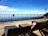 A wooden deck with lounge chairs overlooks a vast scenic view, featuring a bright blue sky with scattered white clouds. A fence runs along the edge of the deck, and there is a lamppost on the right side. The overall ambiance suggests a relaxing outdoor space.