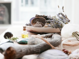 A collection of natural objects including a bundle of lavender, a small bottle of yellow oil, smooth stones, seashells, and driftwood arranged on a light-colored surface. The background is softly blurred, suggesting a peaceful, airy setting.