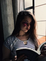 A smiling woman reading a book about saints, seated by a window with natural light.