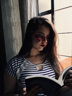 A woman stands by a window, holding an open book in her hands and reading intently. She wears a striped shirt, and her long hair falls over her shoulder. The lighting casts a soft glow on her face, emphasizing a colorful, artistic design around her eye. The atmosphere appears calm and contemplative, with natural light streaming in from outside.