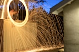 Long exposure photograph capturing a bright circle of sparklers, creating a dynamic swirl of light against a backdrop of trees and a building. The fiery trails are vivid and create an energetic movement across the image.