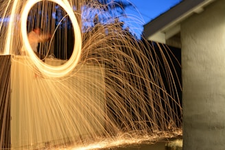 Long exposure photograph capturing a bright circle of sparklers, creating a dynamic swirl of light against a backdrop of trees and a building. The fiery trails are vivid and create an energetic movement across the image.
