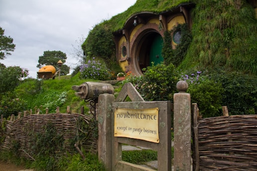 A charming hillside dwelling with a circular, hobbit-like door surrounded by lush green vegetation and vibrant flowers. A wooden sign in the foreground reads 'no admittance except on party business', adding a whimsical touch.