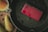 A red handheld gaming console lies on a dark pan surrounded by scattered raisins. A mason jar filled with a yellowish liquid sits nearby on a cork coaster, next to two ripe pears.
