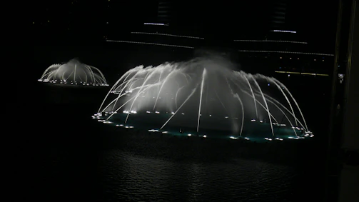 Nighttime scene of a water fountain show illuminated with turquoise and deep blue lights.