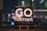 An inspirational message displayed on a small easel features bold white text that reads 'GO FURTHER.' against a cosmic background full of stars and galaxies. The scene is set indoors with a blurred night cityscape in the background, offering colorful bokeh effect lights.
