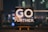 An inspirational message displayed on a small easel features bold white text that reads 'GO FURTHER.' against a cosmic background full of stars and galaxies. The scene is set indoors with a blurred night cityscape in the background, offering colorful bokeh effect lights.