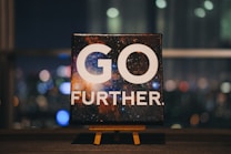 An inspirational message displayed on a small easel features bold white text that reads 'GO FURTHER.' against a cosmic background full of stars and galaxies. The scene is set indoors with a blurred night cityscape in the background, offering colorful bokeh effect lights.