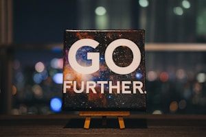 An inspirational message displayed on a small easel features bold white text that reads 'GO FURTHER.' against a cosmic background full of stars and galaxies. The scene is set indoors with a blurred night cityscape in the background, offering colorful bokeh effect lights.