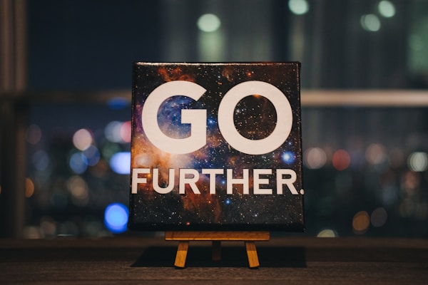 An inspirational message displayed on a small easel features bold white text that reads 'GO FURTHER.' against a cosmic background full of stars and galaxies. The scene is set indoors with a blurred night cityscape in the background, offering colorful bokeh effect lights.