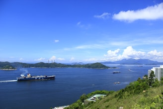 A large cargo ship sails through a wide body of calm water, surrounded by several islands with lush greenery. The sky is clear with some fluffy clouds near the horizon. Some high-rise buildings are located near the shore, and the water reflects the deep blue of the sky.