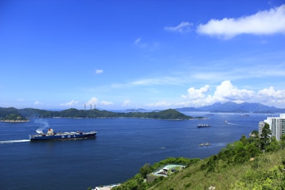 A large cargo ship sails through a wide body of calm water, surrounded by several islands with lush greenery. The sky is clear with some fluffy clouds near the horizon. Some high-rise buildings are located near the shore, and the water reflects the deep blue of the sky.