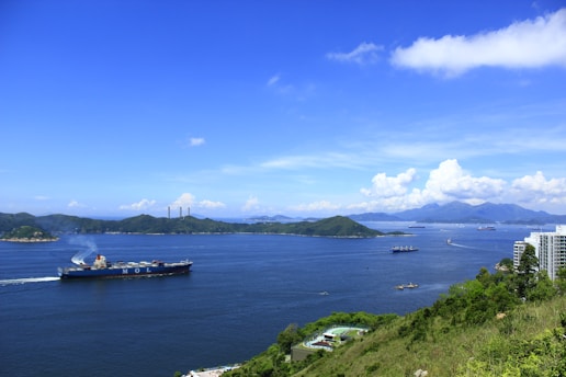 A large cargo ship sails through a wide body of calm water, surrounded by several islands with lush greenery. The sky is clear with some fluffy clouds near the horizon. Some high-rise buildings are located near the shore, and the water reflects the deep blue of the sky.