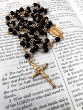A rosary with black beads and gold chain lies on an open Bible, displaying passages from the Book of Psalms. The rosary features a gold crucifix and a medallion as part of its design.