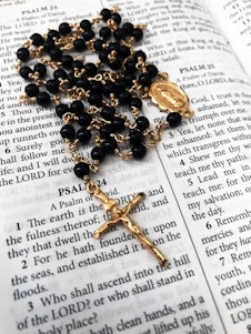 A rosary with black beads and gold chain lies on an open Bible, displaying passages from the Book of Psalms. The rosary features a gold crucifix and a medallion as part of its design.