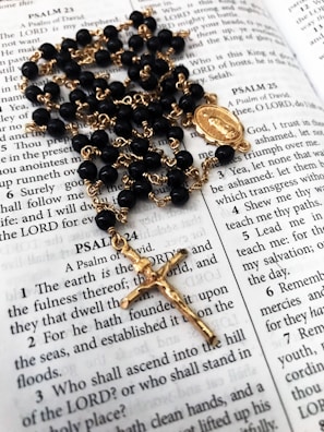A rosary with black beads and gold chain lies on an open Bible, displaying passages from the Book of Psalms. The rosary features a gold crucifix and a medallion as part of its design.