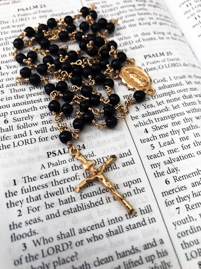 A rosary with black beads and gold chain lies on an open Bible, displaying passages from the Book of Psalms. The rosary features a gold crucifix and a medallion as part of its design.