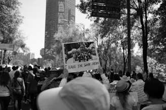 A group of people participating in a street march or protest. One individual prominently holds up a sign with a historical figure giving a speech, captioned 'I HAVE THE SAME DREAM.' The scene is set outdoors with trees and buildings in the background.