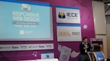A speaker is presenting at a technology-themed event, with a backdrop displaying a title about responsive web design. The backdrop is colorful with purple and blue tones, featuring multiple logos, including IECE, CIESPAL, and Campus Party. The speaker stands at a podium with a microphone, and there is a partial view of the audience area.