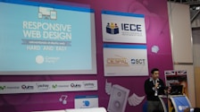 A speaker is presenting at a technology-themed event, with a backdrop displaying a title about responsive web design. The backdrop is colorful with purple and blue tones, featuring multiple logos, including IECE, CIESPAL, and Campus Party. The speaker stands at a podium with a microphone, and there is a partial view of the audience area.