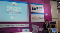 A speaker is presenting at a technology-themed event, with a backdrop displaying a title about responsive web design. The backdrop is colorful with purple and blue tones, featuring multiple logos, including IECE, CIESPAL, and Campus Party. The speaker stands at a podium with a microphone, and there is a partial view of the audience area.
