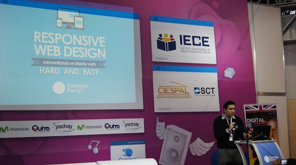 A speaker is presenting at a technology-themed event, with a backdrop displaying a title about responsive web design. The backdrop is colorful with purple and blue tones, featuring multiple logos, including IECE, CIESPAL, and Campus Party. The speaker stands at a podium with a microphone, and there is a partial view of the audience area.