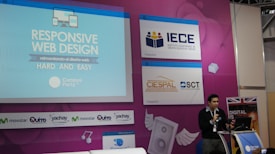 A speaker is presenting at a technology-themed event, with a backdrop displaying a title about responsive web design. The backdrop is colorful with purple and blue tones, featuring multiple logos, including IECE, CIESPAL, and Campus Party. The speaker stands at a podium with a microphone, and there is a partial view of the audience area.