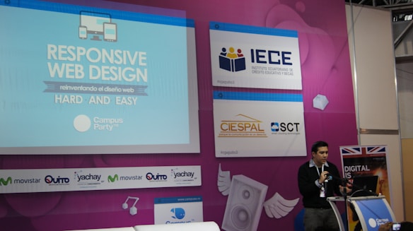 A speaker is presenting at a technology-themed event, with a backdrop displaying a title about responsive web design. The backdrop is colorful with purple and blue tones, featuring multiple logos, including IECE, CIESPAL, and Campus Party. The speaker stands at a podium with a microphone, and there is a partial view of the audience area.