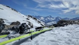 A pair of sturdy, well-used alpine skis resting against a snowy mountain backdrop under a clear blue sky.