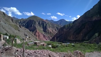 A mountainous landscape with rugged cliffs and multiple shades of green, brown, and red in the rock formations. A small village with simple buildings is nestled at the base of the mountains, surrounded by green vegetation and rocky terrain. The sky is clear with a few scattered clouds, adding depth to the vibrant scenery.