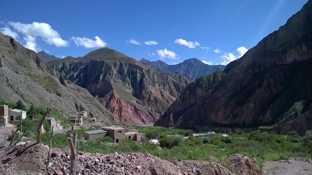 A mountainous landscape with rugged cliffs and multiple shades of green, brown, and red in the rock formations. A small village with simple buildings is nestled at the base of the mountains, surrounded by green vegetation and rocky terrain. The sky is clear with a few scattered clouds, adding depth to the vibrant scenery.