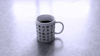 A white coffee mug printed with various black geometric and abstract symbols sitting on a light gray surface.