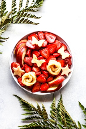 A bowl of fresh fruit including sliced strawberries, apple slices, and decorative shapes of star and butterfly cutouts made from apple. The setup is complemented by green fern leaves arranged around the bowl on a light background.